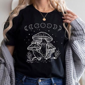 May include: Black t-shirt with a white graphic of mushrooms and a crescent moon phase design. The text "Shroom Queens" is printed on the shirt.