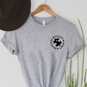 May include: Gray t-shirt with a black puzzle piece graphic and the words "Accept, Adapt, Advocate" around it.
