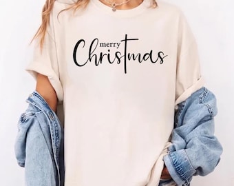 May include: A cream-colored short-sleeve t-shirt featuring the black text Merry Christmas printed on the front, with the letter t stylized as a cross. The shirt is worn with a light blue denim jacket and denim shorts, creating a casual look.
