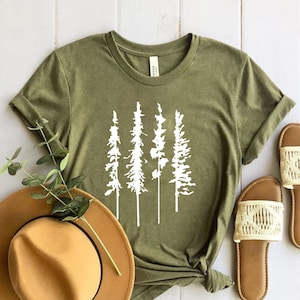 May include: A green t-shirt with a white graphic of four trees. The t-shirt is laid out on a white wooden surface with a brown hat and two pairs of brown sandals with white straps.