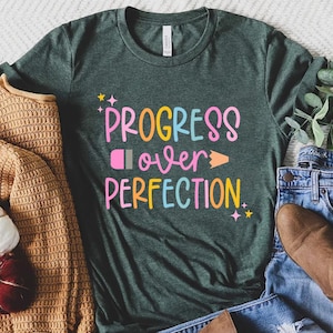 May include: A dark green t-shirt with the text "PROGRESS over PERFECTION" in colorful letters. The shirt is styled with a brown sweater, blue jeans, and brown ankle boots.