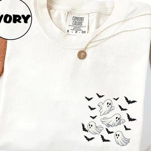 May include: Ivory-colored t-shirt featuring a graphic of cartoon ghosts and black bats. The shirt has a crew neck and short sleeves. A gold necklace with a pendant is draped across the shirt. The shirt is a Comfort Colors brand.