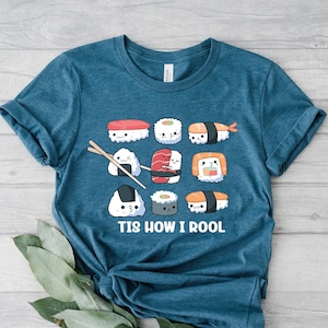 May include: Teal t-shirt featuring a sushi-themed graphic. The design includes various sushi rolls with cute faces and chopsticks. The text "Tis How I Rool" is printed below the sushi illustrations. The shirt is made of a soft material.