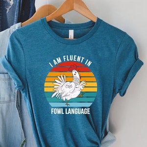 May include: Teal t-shirt with a graphic of a white chicken inside a sunset circle. The text reads "I AM FLUENT IN FOWL LANGUAGE". The shirt is hanging on a wooden hanger with other clothing.