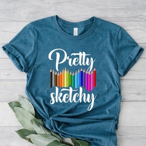 以下が含まれることがあります： ターコイズブルーのTシャツに、白いグラフィックデザインで「Pretty Sketchy」と書いてあり、その下にカラフルな鉛筆が並んでいます。