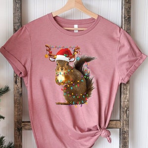 May include: A pink t-shirt with a graphic of a squirrel wearing a Santa hat and reindeer antlers. The squirrel is holding a string of Christmas lights.