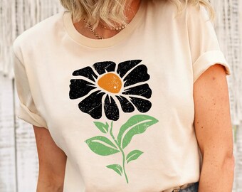 May include: Cream-colored t-shirt featuring a large, stylized flower graphic. The flower has black petals with an orange center, green leaves, and a green stem. The shirt has rolled-up sleeves and a crew neck.