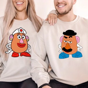 May include: A couple wearing matching cream-colored sweatshirts with cartoon images of Mr. and Mrs. Potato Head. The woman's sweatshirt features Mrs. Potato Head with a pink nose and a white flower on her head. The man's sweatshirt features Mr. Potato Head with a black hat and a brown mustache.