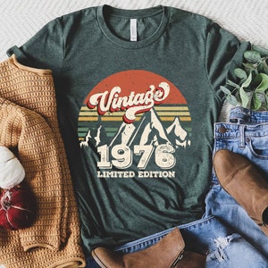 May include: A dark green t-shirt with a vintage 1976 design featuring a sunset, mountains, and the text "Vintage 1976 Limited Edition".