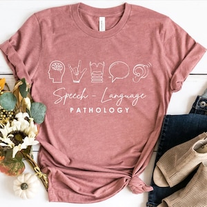 May include: A dusty rose t-shirt with white line art illustrations and the words "Speech-Language Pathology". The design includes a brain, hand gestures, a throat, a speech bubble, and an ear. The shirt is folded and styled with jeans and sandals.