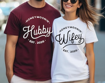 May include: A maroon t-shirt with white text reading HONEYMOONIN' Hubby EST. 2026 and a white t-shirt with black text reading HONEYMOONIN' Wifey EST. 2026. Both shirts feature short sleeves, crew necklines, and a graphic design centered on the chest area.