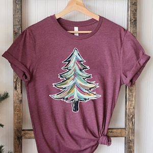 May include: A maroon t-shirt with a colorful abstract Christmas tree design. The tree is painted in shades of green, blue, yellow, and red.