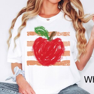 May include: A white short-sleeve t-shirt featuring a graphic of a red apple with a green leaf, rendered in a textured, scribbled crayon style. The apple is layered over four horizontal, distressed peach-colored stripes across the center.