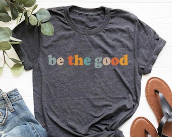 May include: Dark gray t-shirt with the text "be the good" in colorful letters. The shirt is paired with blue jeans and brown sandals with black straps. A sprig of greenery is in the background.
