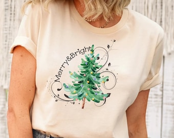 May include: Cream-colored t-shirt featuring a watercolor Christmas tree design with the words "Merry & Bright" in a script font. The tree is green with colorful lights and a brown trunk, surrounded by decorative swirls.