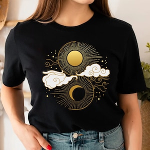 May include: Black t-shirt with a gold sun and moon design. The design features stylized clouds and stars.