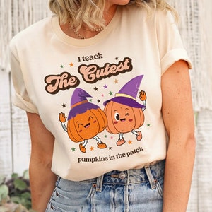 May include: Cream-colored t-shirt featuring the text "I teach the Cutest" above two cartoon pumpkins wearing witch hats. The pumpkins are orange with smiling faces and are dancing. The text "pumpkins in the patch" is below the pumpkins.