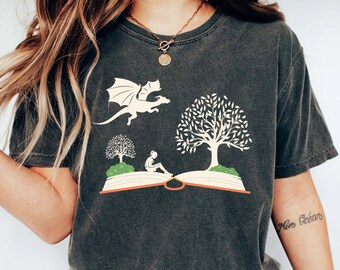 May include: A dark gray t-shirt with a graphic of a dragon flying over an open book with a person sitting on the book. The book is surrounded by two trees. The text "Mon Océan" is visible on the person's arm.