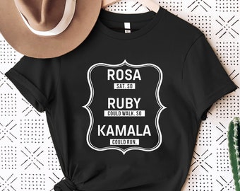 May include: Black t-shirt with a white graphic design featuring the names Rosa, Ruby, and Kamala with quotes about their actions. The text reads "Rosa sat. So Ruby could walk. So Kamala could run."