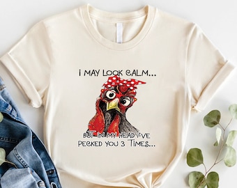 May include: A beige t-shirt with a graphic of a cartoon chicken wearing red glasses and a red bandana with white polka dots. The text on the shirt reads "I may look calm... But in my head I've pecked you 3 times..."