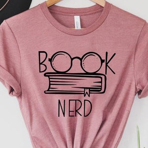 May include: Mauve t-shirt with the words "BOOK NERD" in black, along with a graphic of glasses over a stack of books. The shirt is hanging on a wooden hanger against a white background. A black hat is visible in the background.