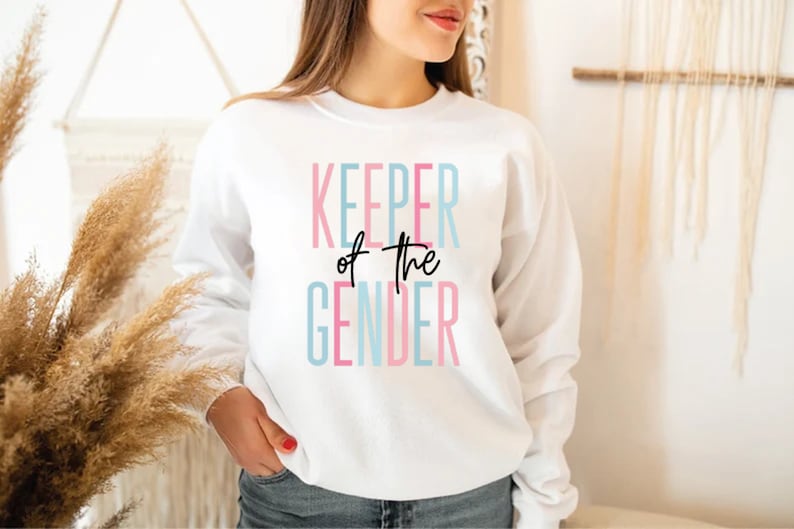 May include: White sweatshirt with the text "KEEPER of the GENDER" in pink, blue, and black lettering.