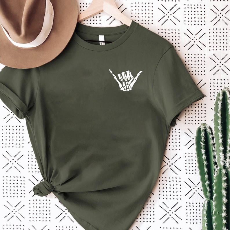 Op de afbeelding: Een groen T-shirt met korte mouwen met een witte skelethand die een shaka-teken maakt.