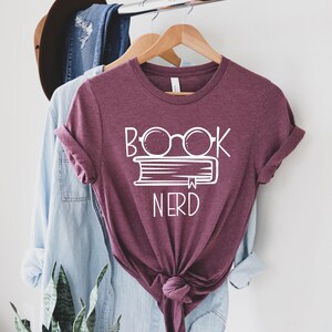 May include: A maroon t-shirt with the words "BOOK NERD" in white, featuring glasses over a book. The shirt is hanging on a wooden hanger with other clothing items in the background. The shirt is knotted at the bottom.