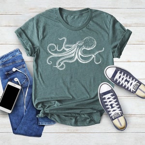 May include: A teal t-shirt featuring a white octopus illustration. The shirt is paired with blue jeans, a smartphone with white earbuds, and navy blue sneakers with white laces. The items are arranged on a light wood-grain surface.