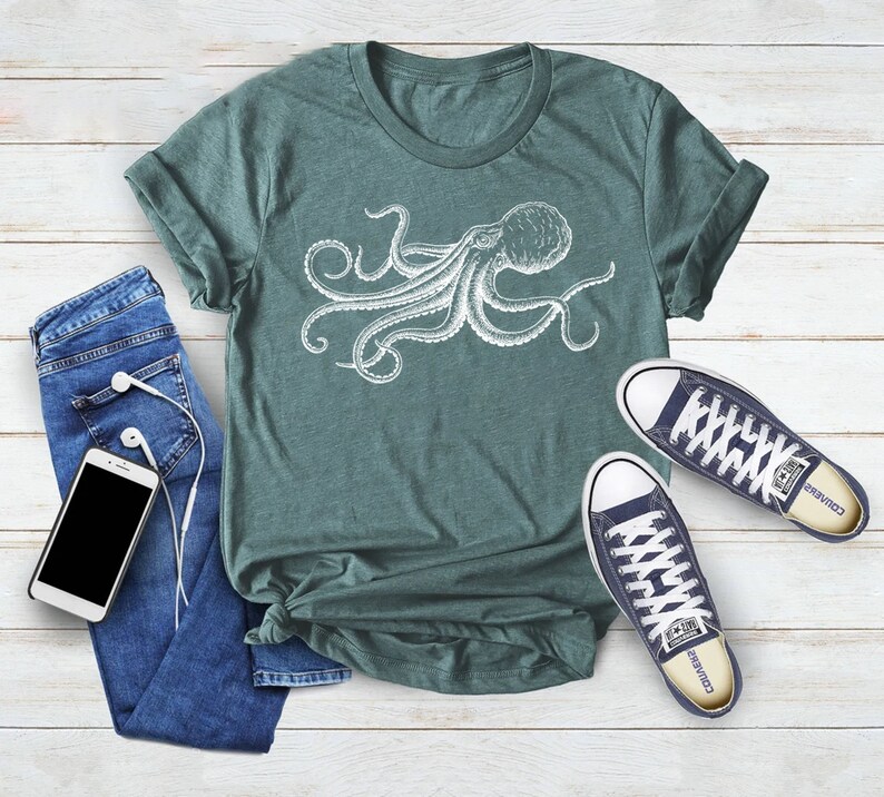 May include: A teal t-shirt featuring a white octopus illustration. The shirt is paired with blue jeans, a smartphone with white earbuds, and navy blue sneakers with white laces. The items are arranged on a light wood-grain surface.