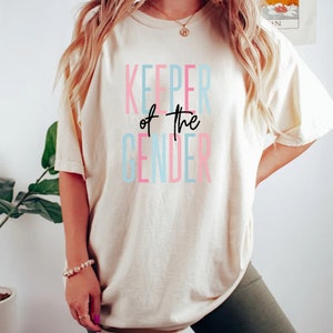 May include: A white t-shirt with the text "KEEPER of the GENDER" in pink, blue, and black lettering.