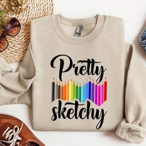 以下が含まれることがあります： ベージュのトレーナーに、黒い文字で「Pretty sketchy」と書いてあります。テキストの上に、カラフルな鉛筆が並んでいます。