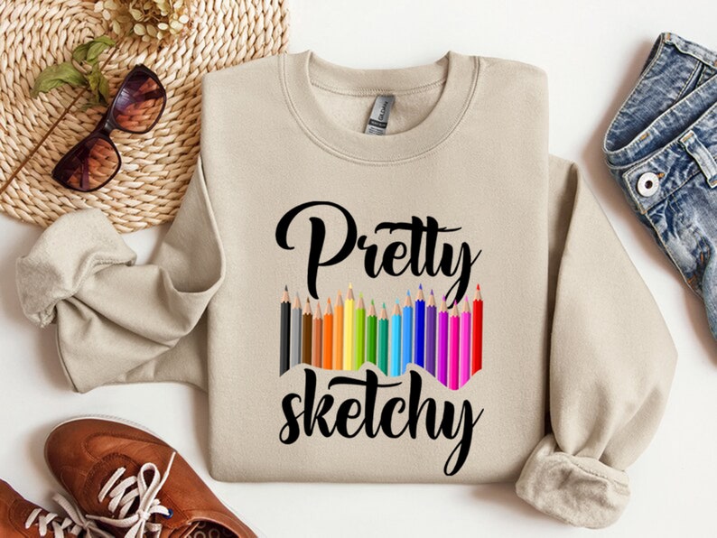 以下が含まれることがあります： ベージュのトレーナーに、黒い文字で「Pretty sketchy」と書いてあります。テキストの上に、カラフルな鉛筆が並んでいます。