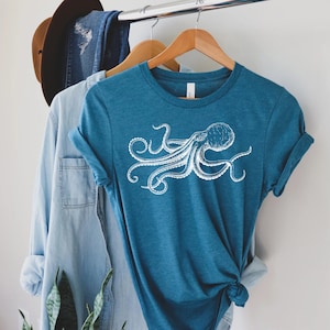 May include: Teal-colored t-shirt featuring a white octopus illustration. The graphic tee has short sleeves and a crew neck. The shirt is hanging on a wooden hanger with other clothing items, including a denim shirt and a brown hat.