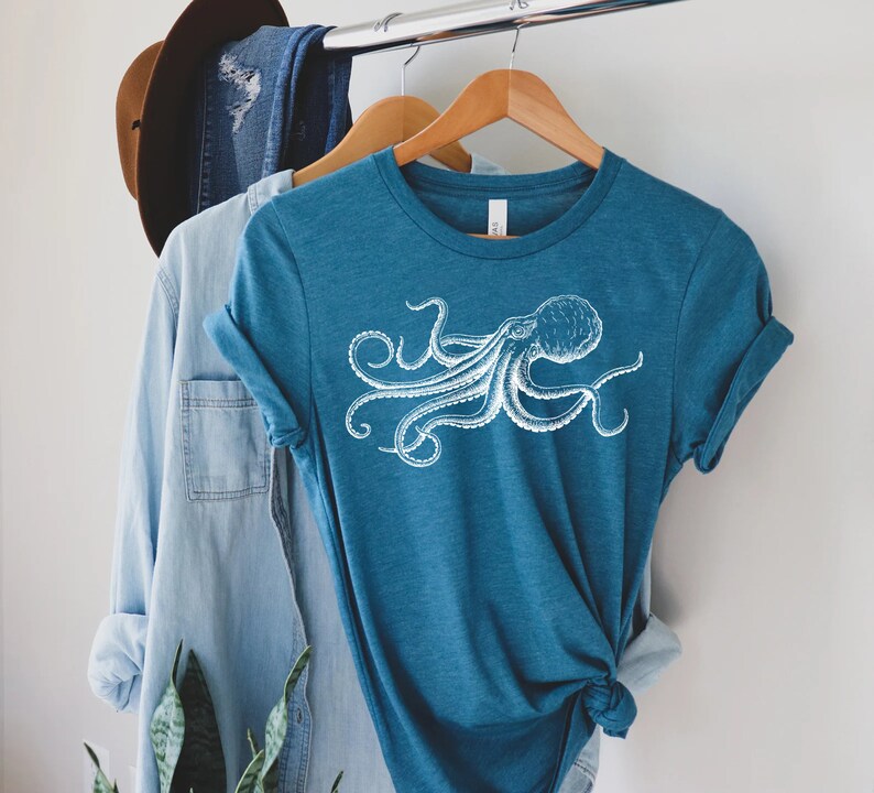 May include: Teal-colored t-shirt featuring a white octopus illustration. The graphic tee has short sleeves and a crew neck. The shirt is hanging on a wooden hanger with other clothing items, including a denim shirt and a brown hat.