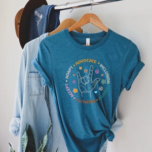 May include: A teal blue t-shirt with a circle graphic featuring a hand making the American Sign Language sign for "I love you". The circle graphic also includes the words "Adapt", "Accept", "Equality", "Advocate", "Inclusion", and "Equity" around the hand sign.