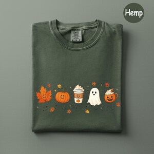 May include: A dark olive green t-shirt featuring a graphic print of a smiling orange maple leaf, a smiling pumpkin, a coffee cup with a pumpkin design, a white ghost, and a jack-o'-lantern mug, all surrounded by small orange and yellow leaves.