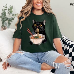 May include: A woman wearing a dark green t-shirt with a black cat eating ramen noodles from a bowl. The cat is wearing a red collar and has a white egg in the bowl. The t-shirt has the word "FOREST" printed on the back.