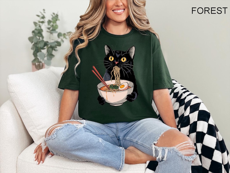 May include: A woman wearing a dark green t-shirt with a black cat eating ramen noodles from a bowl. The cat is wearing a red collar and has a white egg in the bowl. The t-shirt has the word "FOREST" printed on the back.