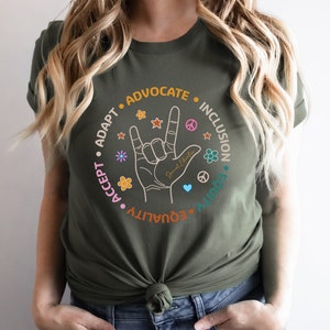 May include: A green t-shirt with a circle graphic featuring the American Sign Language symbol for "I love you" surrounded by the words "Accept", "Adapt", "Equality", "Equity", "Advocate", "Inclusion".