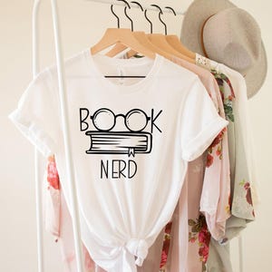 May include: White t-shirt with a black graphic of glasses over a book and the word "NERD" underneath. The shirt is hanging on a wooden hanger with other clothing items, including a floral patterned top and a light-colored hat.