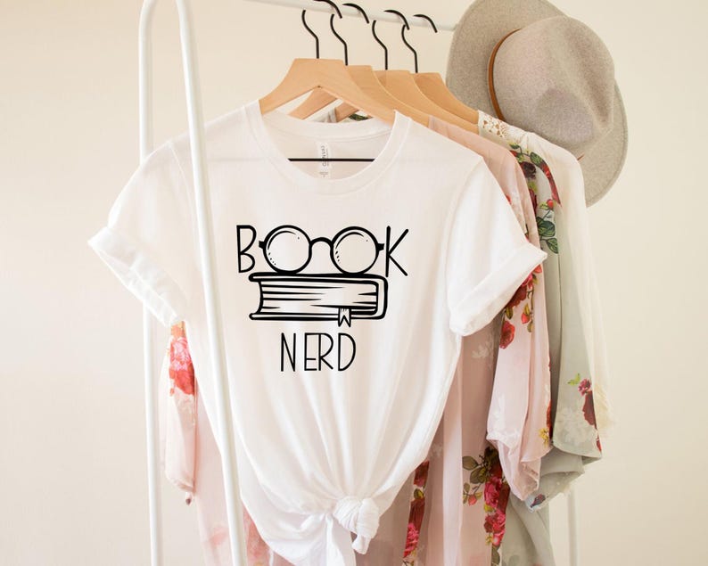 May include: White t-shirt with a black graphic of glasses over a book and the word "NERD" underneath. The shirt is hanging on a wooden hanger with other clothing items, including a floral patterned top and a light-colored hat.