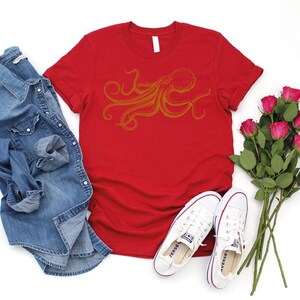 May include: A red t-shirt with a gold octopus design, paired with a denim shirt, white sneakers, and a bouquet of pink roses. The casual ensemble suggests a relaxed, everyday style.