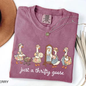 May include: Berry-colored t-shirt with a crew neck. The shirt features a whimsical illustration of geese dressed in vintage clothing, with the text "just a thrifty goose" in a white script. A silver necklace with a pendant is draped across the shirt.
