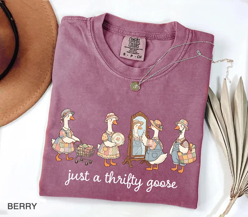 May include: Berry-colored t-shirt with a crew neck. The shirt features a whimsical illustration of geese dressed in vintage clothing, with the text "just a thrifty goose" in a white script. A silver necklace with a pendant is draped across the shirt.