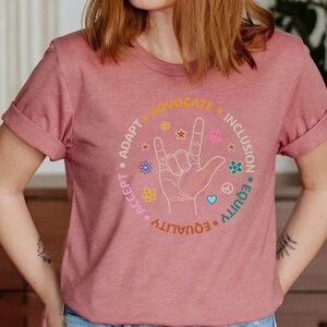 May include: A pink t-shirt with a graphic of a hand signing "I love you" in American Sign Language. The graphic is surrounded by a circle with the words "Accept, Adapt, Advocate, Equality, Equity, Inclusion".