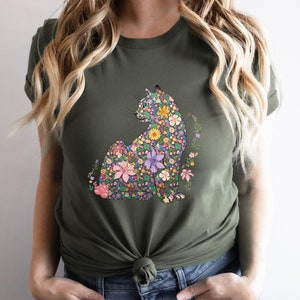 May include: Olive green t-shirt featuring a floral cat design. The cat is composed of various colorful flowers, including pink, yellow, and purple blooms. The shirt has short sleeves and a crew neck.