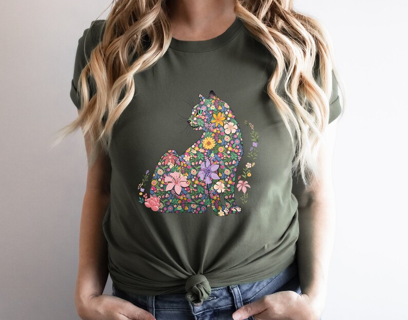 May include: Olive green t-shirt featuring a floral cat design. The cat is composed of various colorful flowers, including pink, yellow, and purple blooms. The shirt has short sleeves and a crew neck.