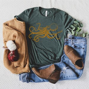 May include: A green t-shirt with an orange octopus design, paired with blue jeans, brown boots, and a tan sweater. A decorative bundle of yarn and a small pumpkin are also present.