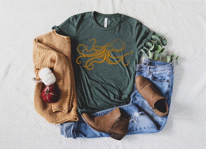 May include: A green t-shirt with an orange octopus design, paired with blue jeans, brown boots, and a tan sweater. A decorative bundle of yarn and a small pumpkin are also present.